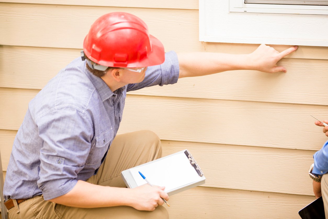 Repairman, building inspector, exterminator, engineer, insurance adjuster, or other blue collar worker has just begun his examination of a building/home's exterior wall, window, and foundation. He wears a red hard hat and clear safety glasses and writes on his clipboard.  Selective focus on blank paper for copy.